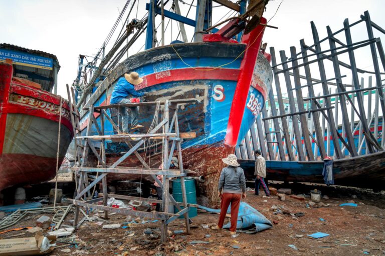 Workers actively repair colorful wooden fishing boats on a dockside, showcasing craftsmanship and teamwork.